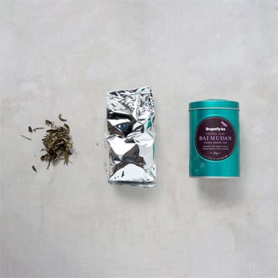 Dragonfly Tea Tin Packaging Layout on a Grey Marble Effect Background. Loose Leaf Bai Mudan White Tea Leaves, the Inner Pouch and Outer Tin.